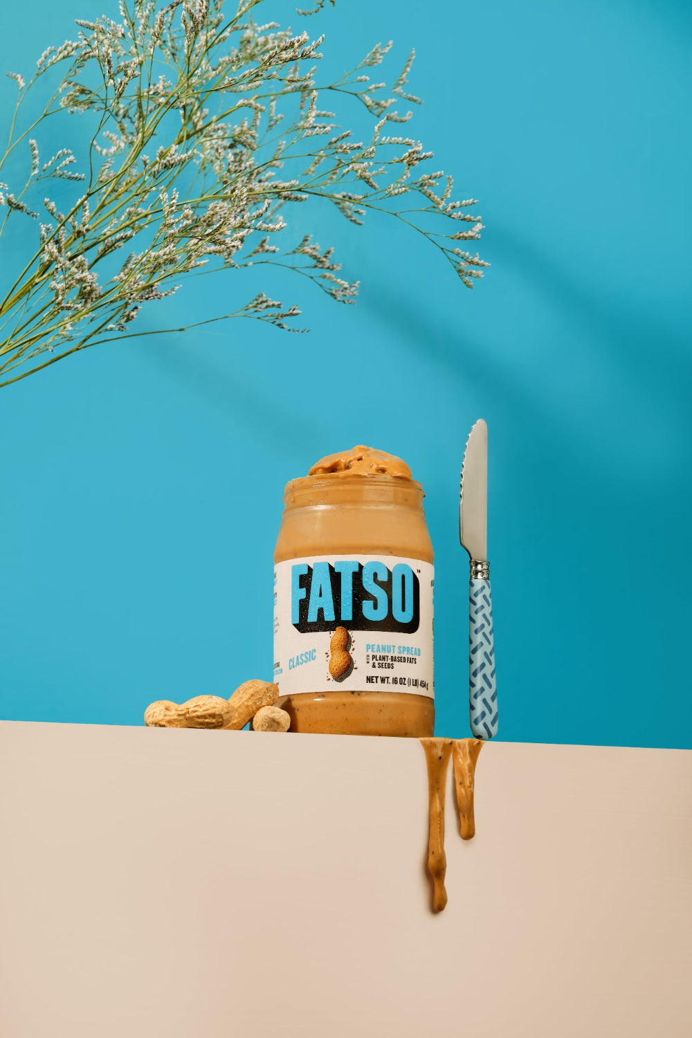 A jar of FATSO Peanut Butter in the Peanut Butter Classic flavor, styled on a beige surface with peanut butter dripping down, a patterned knife, peanuts, and dried flowers against a bright blue background.