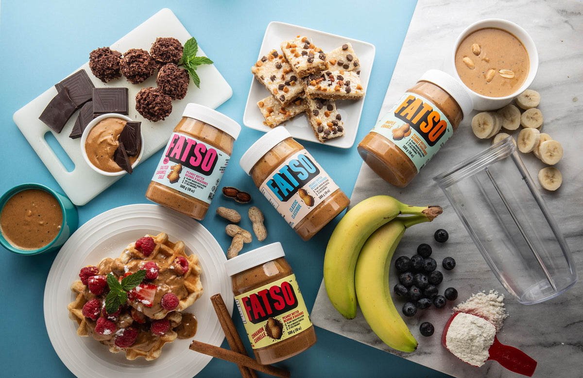 Three jars of Fatso peanut butter arranged on a light blue surface with various food items including chocolate-covered dates, chocolate chips waffles, banana slices, blueberries, and a clear blender jar.
